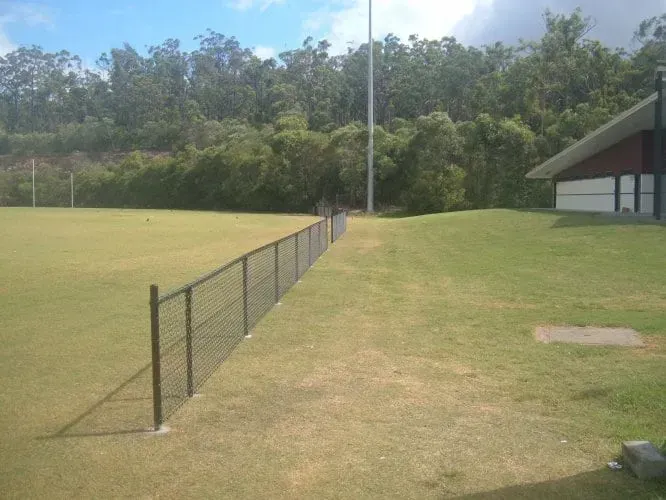 A wire fence separates a green grass sports field from a grassy path next to a building, with trees in the background.