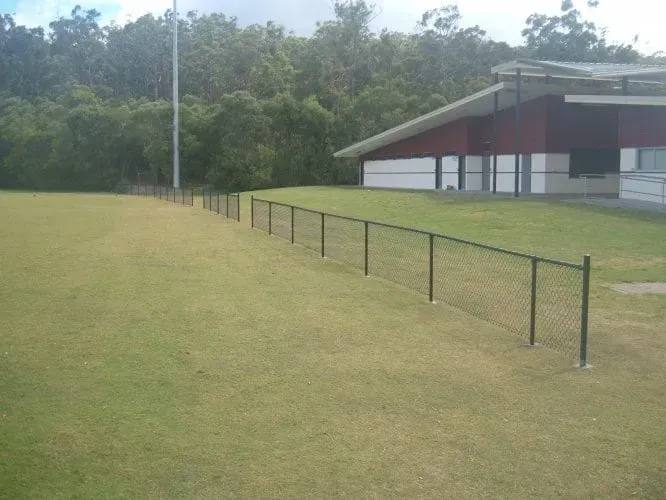 A chain-link fence divides a grassy field from a building with a red and white exterior, bordered by trees.