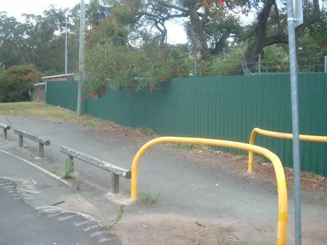 A paved path with wooden railings and yellow metal bollards alongside a high green privacy fence.