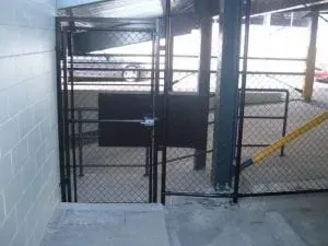 A black metal chain-link gate leading to a concrete parking ramp, viewed from a top-of-stairs landing.