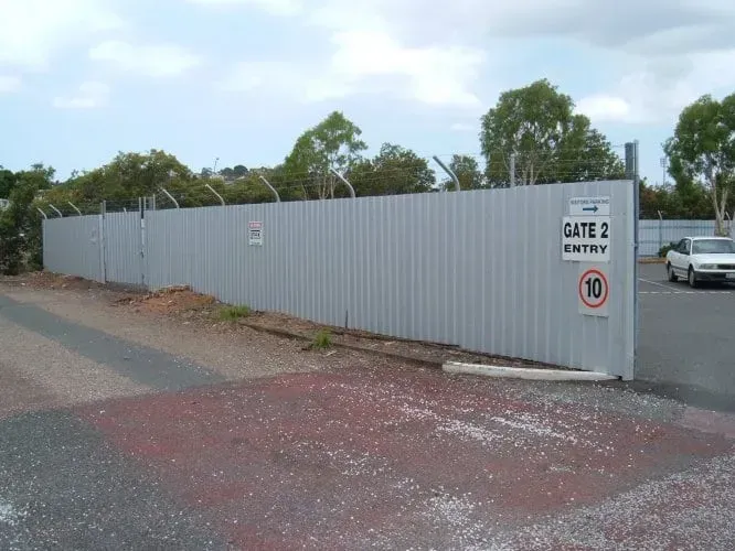A tall, corrugated metal security fence topped with barbed wire, with a sign reading 