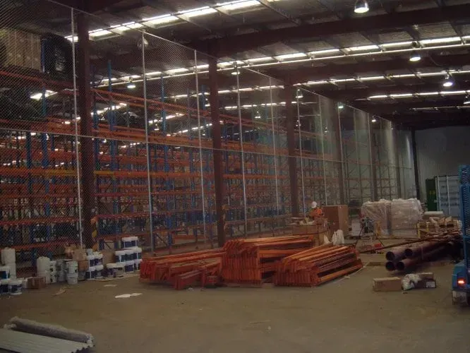 A warehouse interior under construction featuring tall metal shelving units, stacked orange beams, and scattered materials.