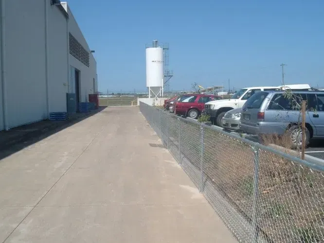 A paved walkway beside a white warehouse building, separated from a parking lot by a chain-link fence on a sunny day.