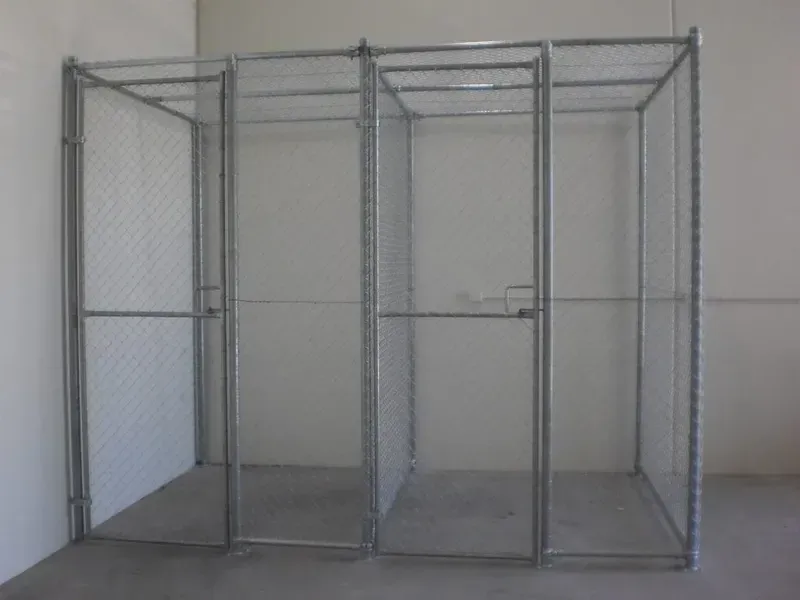 A modular, silver metal chain-link security enclosure or storage cage with two hinged doors, standing against a white wall.