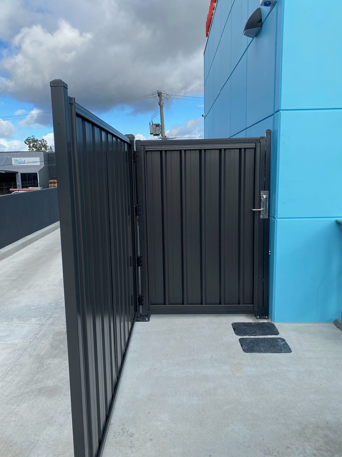 A dark gray metal gate attached to a bright blue building on a concrete surface under a cloudy sky.