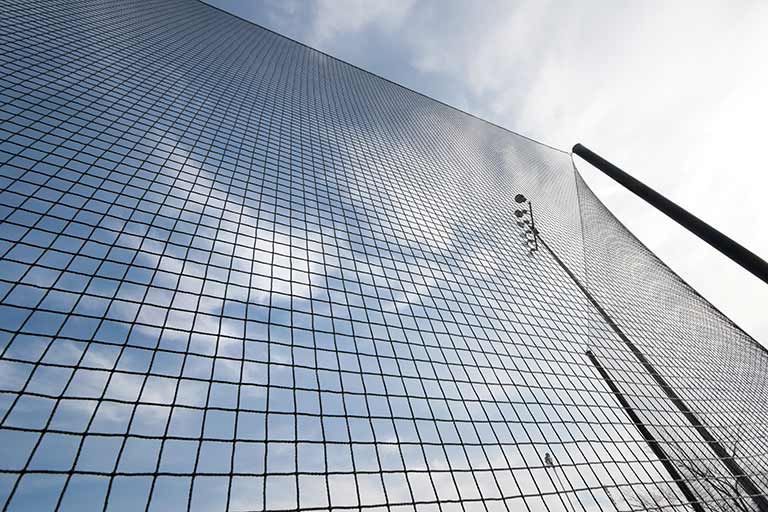 A tall, dark sports netting structure angled against a blue sky with thin clouds.