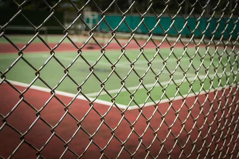 Close-up of a metal chain-link fence in the foreground, with an out-of-focus tennis court visible in the background.