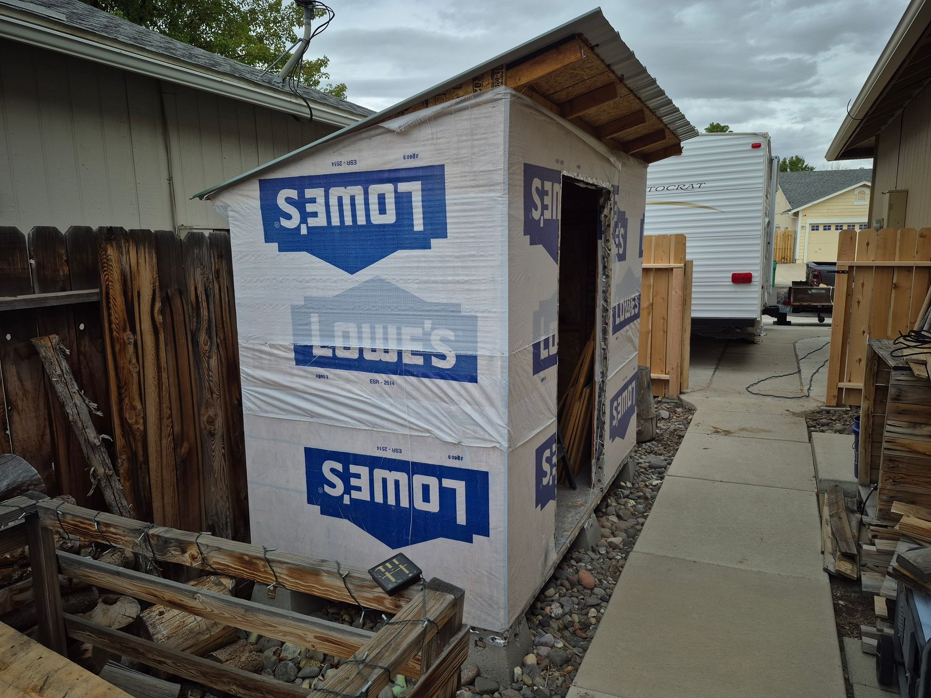 Small shed under construction wrapped in Lowe's logo material, between a wooden fence and a building.