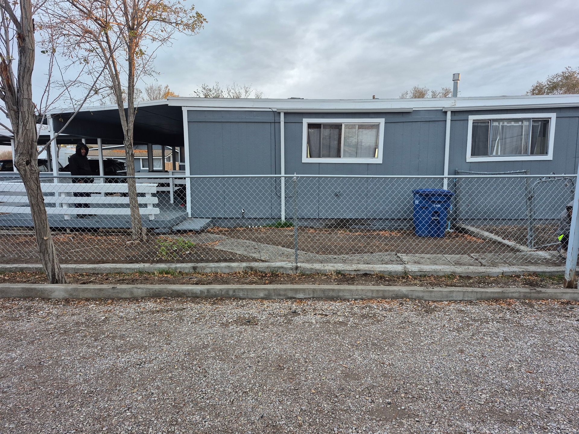 Gray mobile home behind chain-link fence on a gravel surface. Cloudy sky.