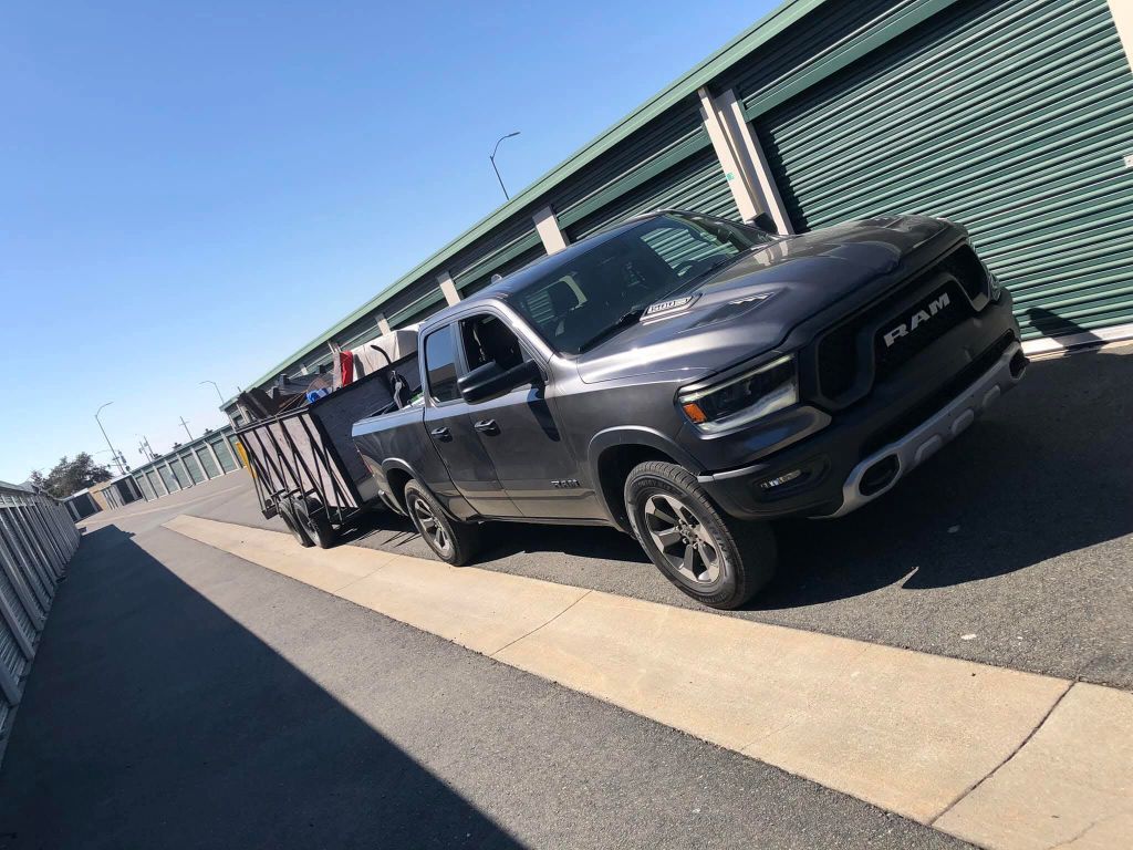 Dark gray Ram truck with a trailer parked in front of storage units on a sunny day.