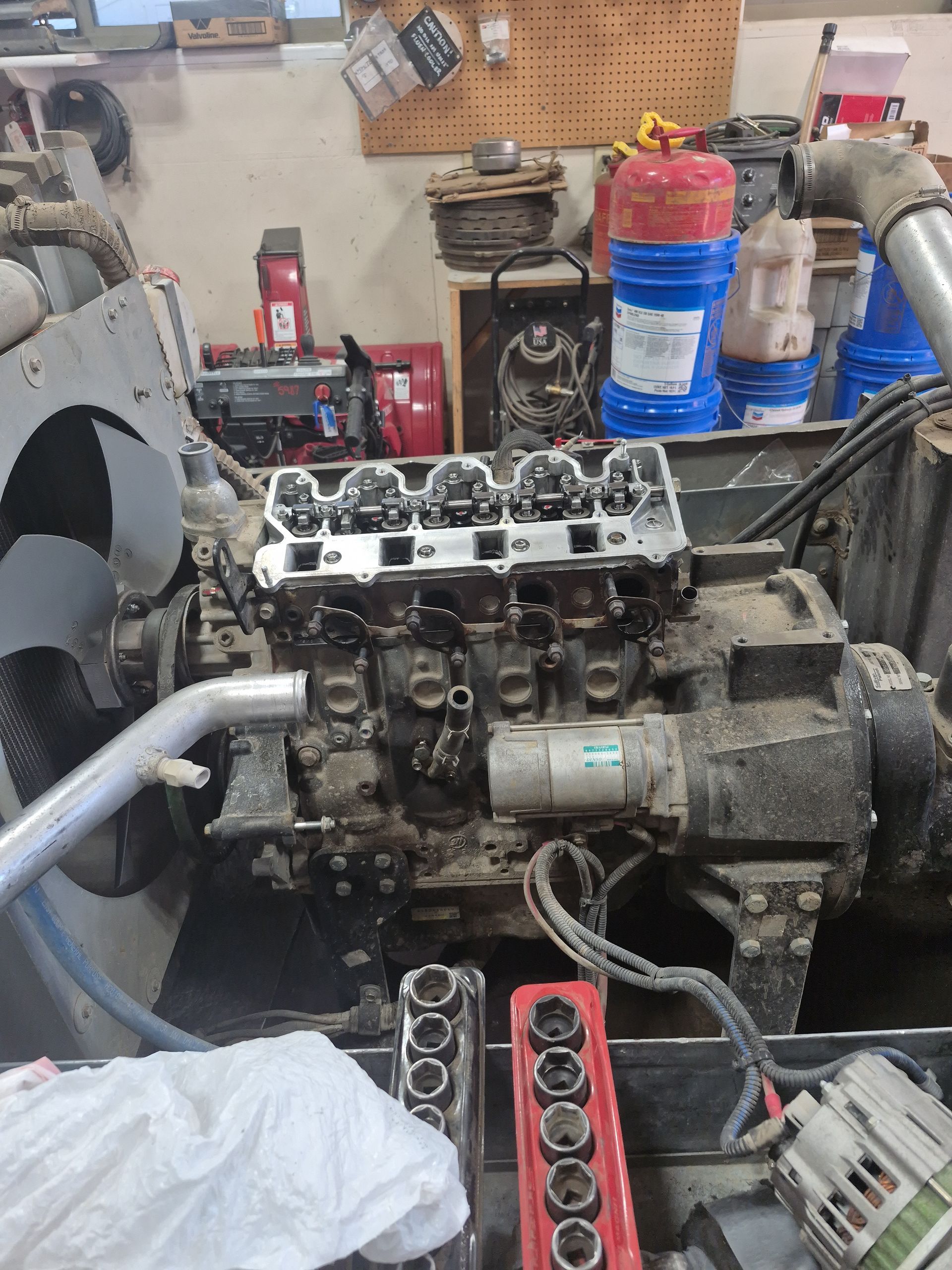 Engine with cylinder head removed in a repair shop. Tools, oil containers, and fan are visible.