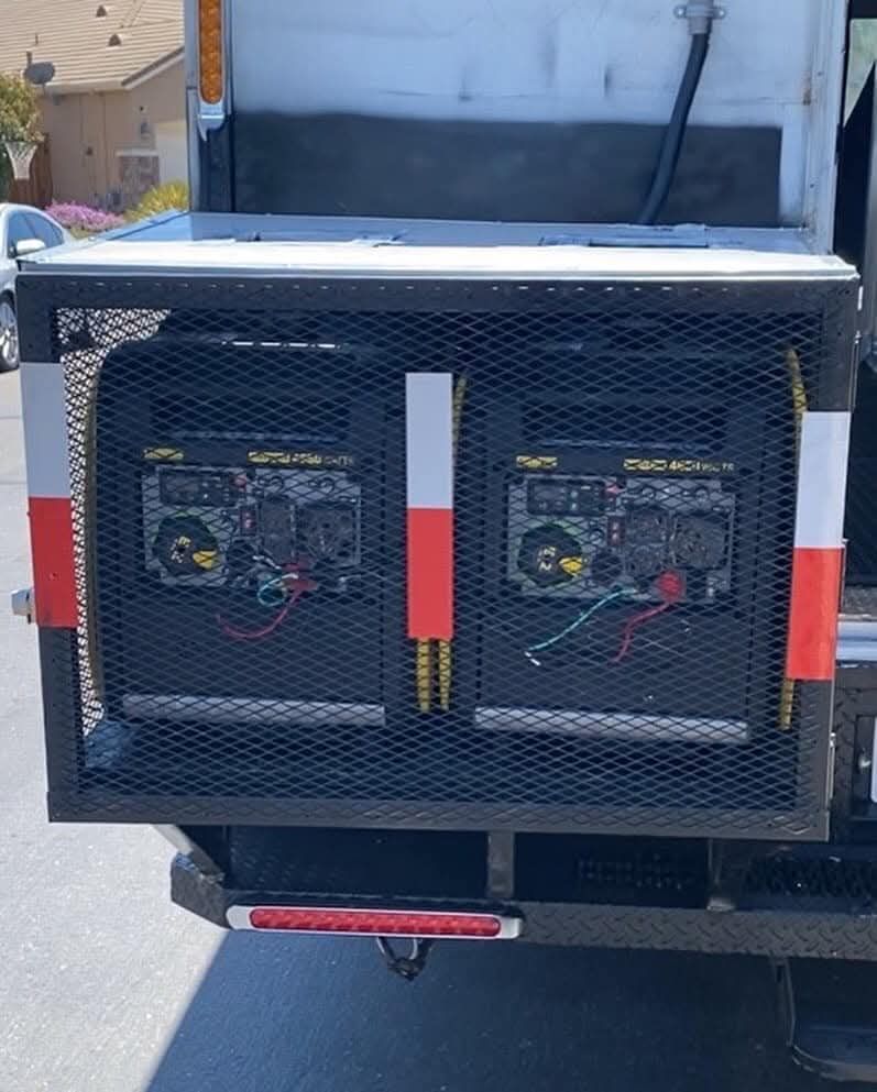 Two generators inside a black metal cage on the back of a truck, with red and white reflective stripes.