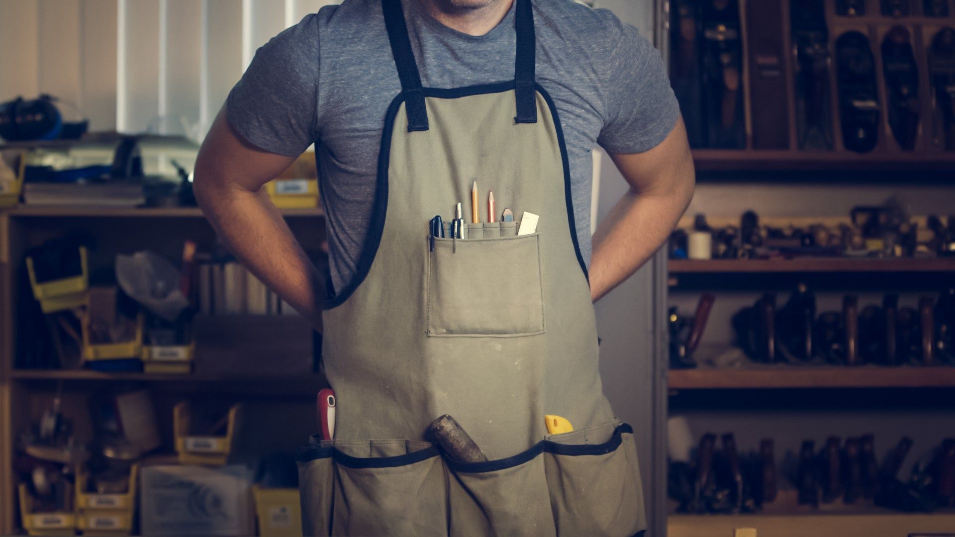 Person wearing a tan apron in a workshop, hands behind their back; tools on shelves.