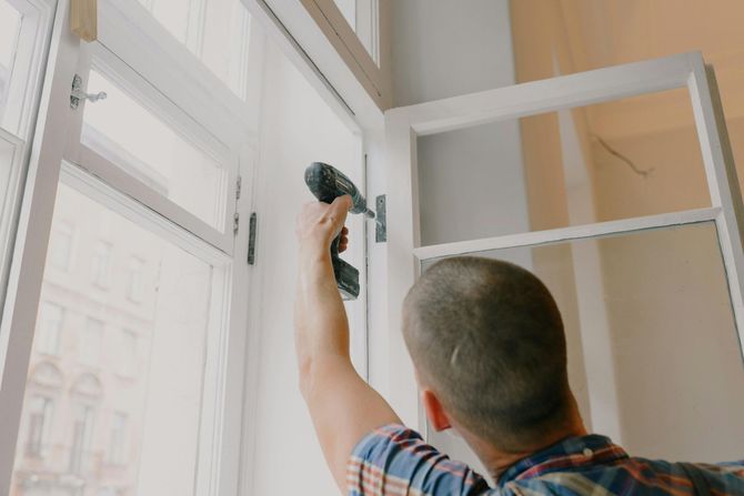 Person using a power drill on a white window frame, indoors.