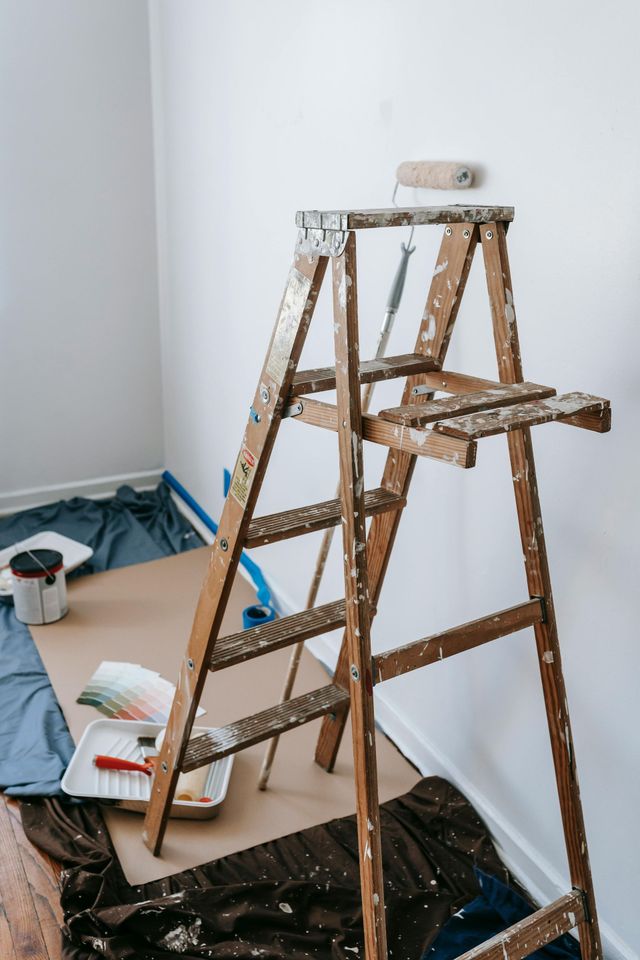 Ladder with paint roller in a room being painted, paint supplies on floor covered with drop cloths.