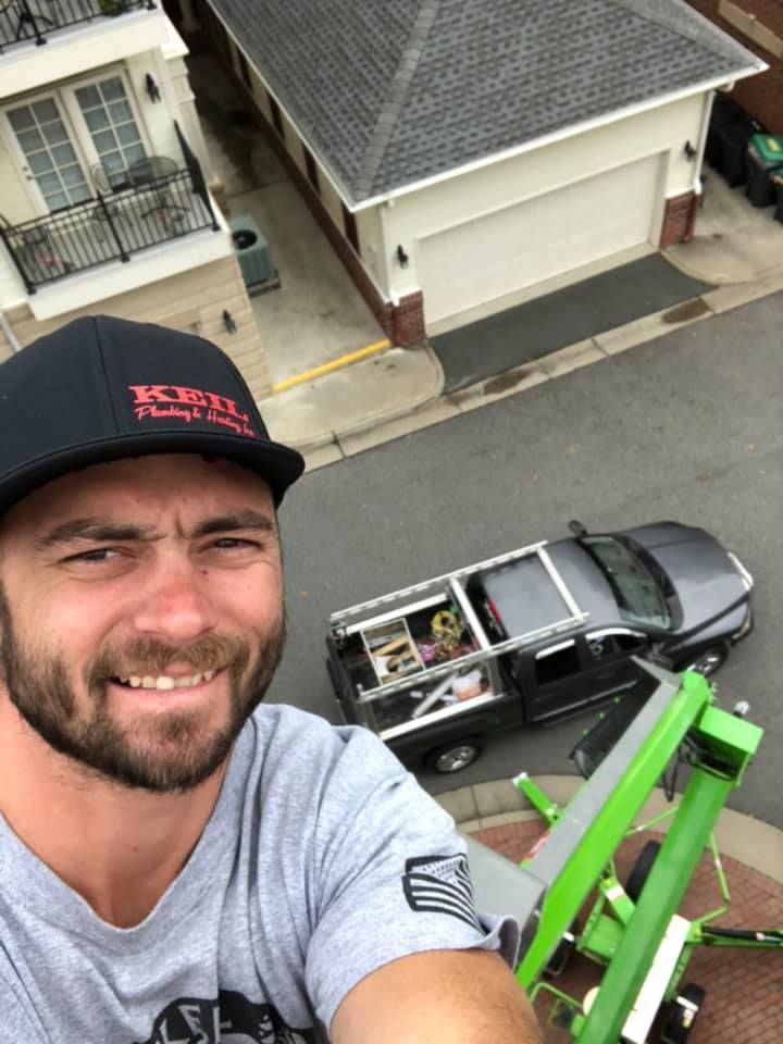 Man in cap smiles, taking a selfie from a lift; truck and buildings in the background.