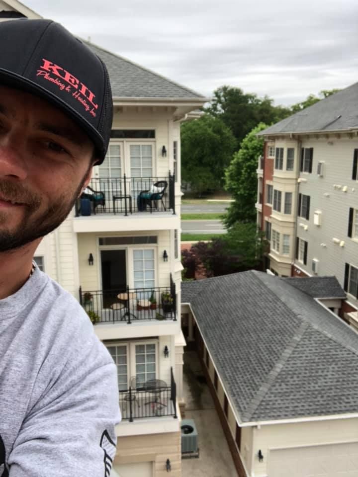 Man in baseball cap takes a selfie, overlooking a multistory apartment building and a dark gray roof.