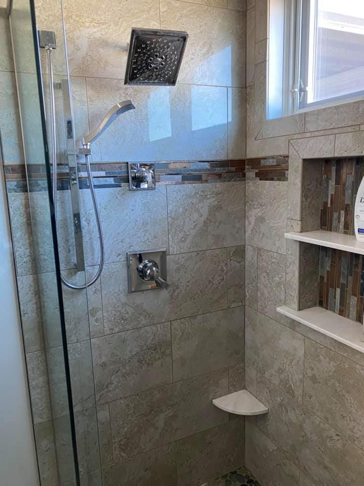 Modern tiled shower with glass door, rain showerhead, handheld, recessed shelves, and corner seat.