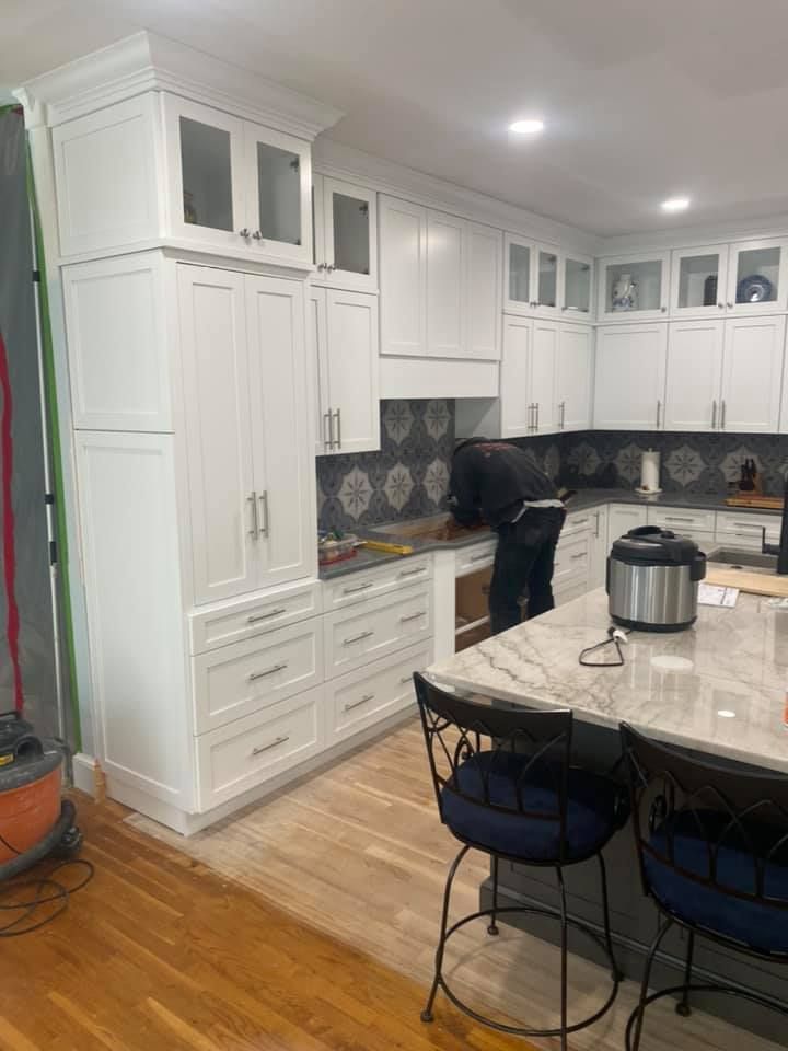 A person works in a bright white kitchen with wooden floors, installing cabinets.
