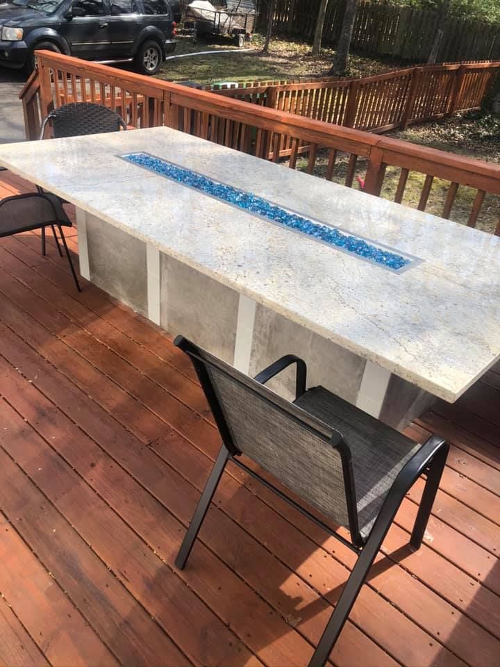 Outdoor rectangular fire pit table on a wooden deck with chairs; blue fire glass.