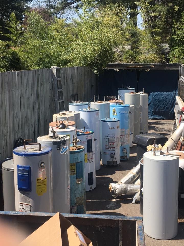 Row of discarded water heaters, mostly white and blue, in a yard near a fence.