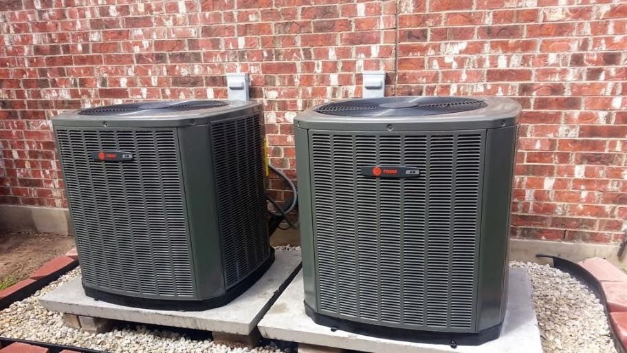 Two dark HVAC units sit on concrete pads against a brick wall.