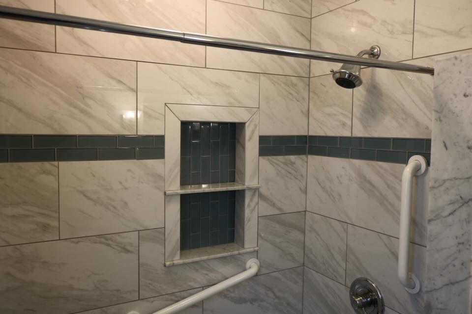 Bathroom shower with marble tile, grab bars, built-in shelf, and chrome shower head.