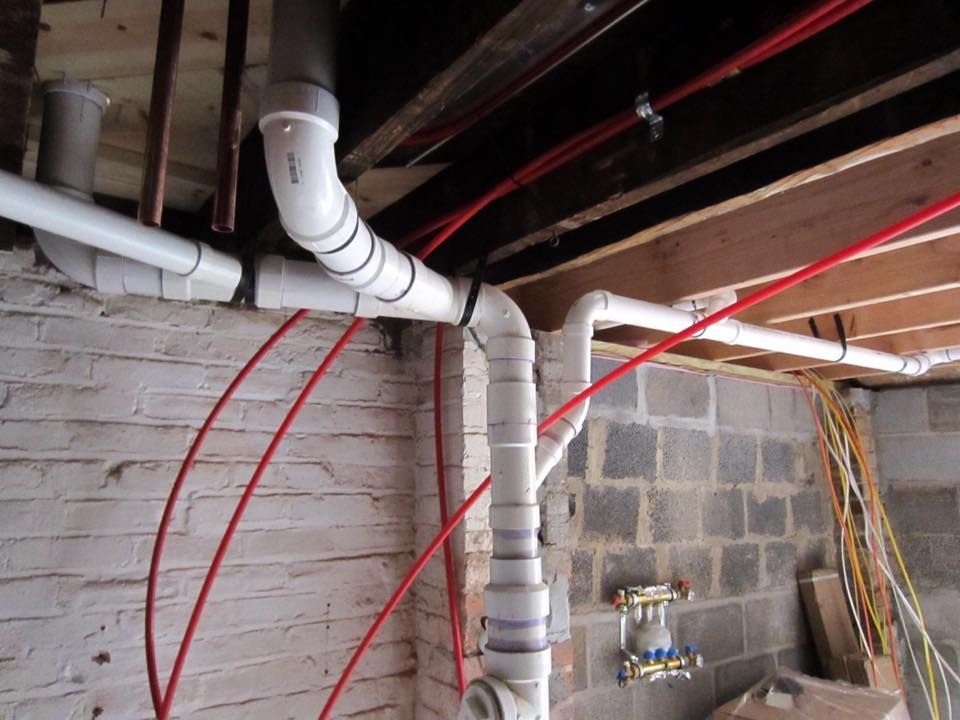 White PVC pipes and red tubing in a basement; pipes are connected, with some near brick and a wooden ceiling.