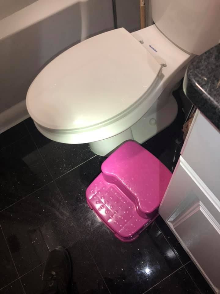 Pink toilet step stool in front of a white toilet on black tile flooring in a bathroom.