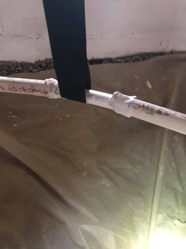 White PVC pipe with patched connections, secured by black strap, in a crawl space.