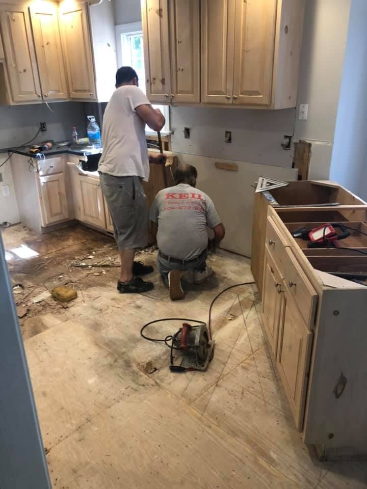 Two workers remodel a kitchen, one using a tool, the other standing. Cabinets and debris are visible.