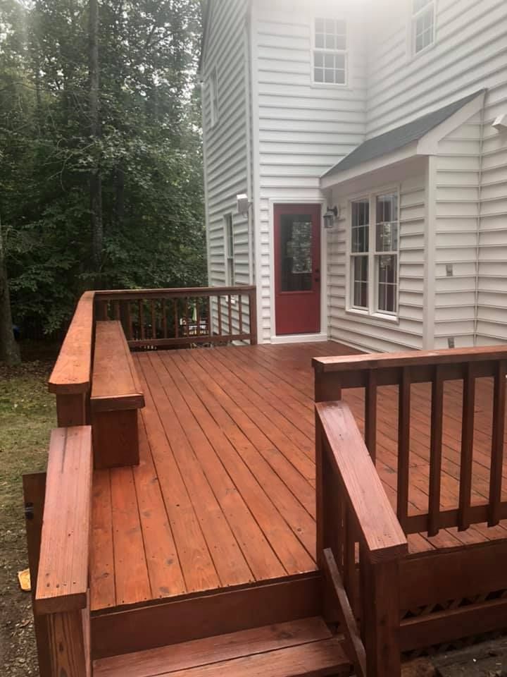 Wooden deck with red-stained wood, stairs, and benches next to a two-story white house with a red door.