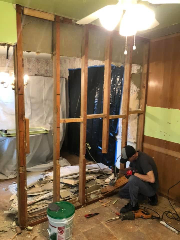 Man demolishes a wall, wood frame exposed. Green and brown walls surround. Debris litters floor, green bucket visible.