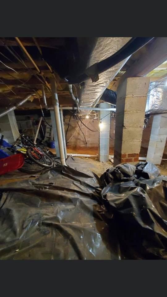 Crawl space with plastic sheeting on the ground, supporting beams, pipes, and a cluttered area with various items.