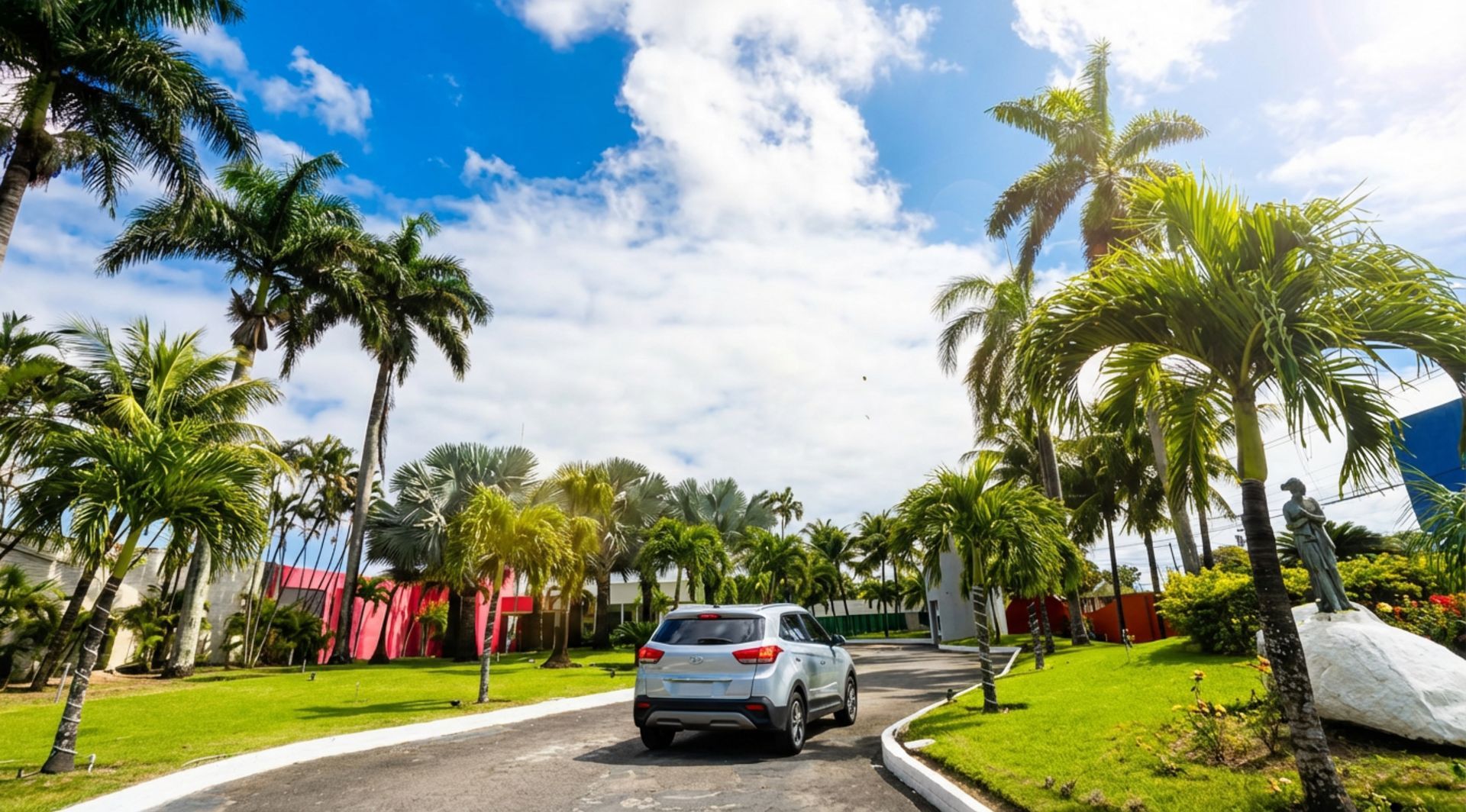 Entrada de automóveis que leva a uma propriedade murada com palmeiras sob um céu azul.
