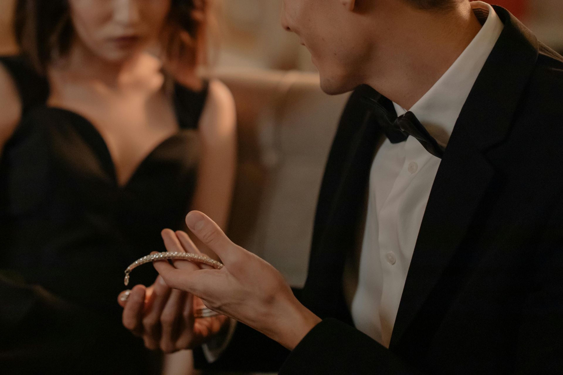 Homem de smoking presenteia uma mulher de vestido preto com uma pulseira de diamantes.