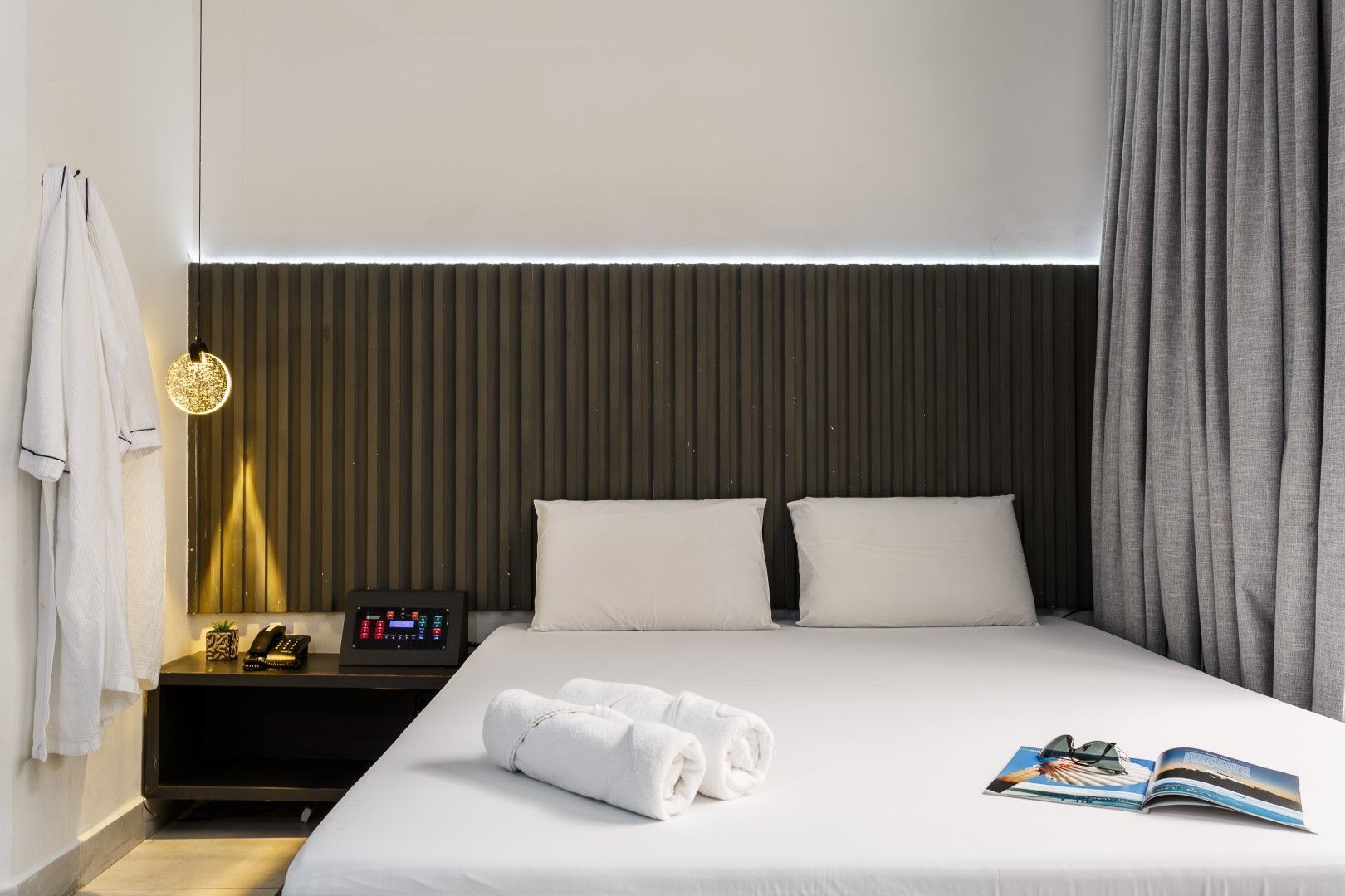 Quarto com cama, criado-mudo, roupão e cortinas. Cabeceira marrom-escura. Lençóis e toalhas brancos.