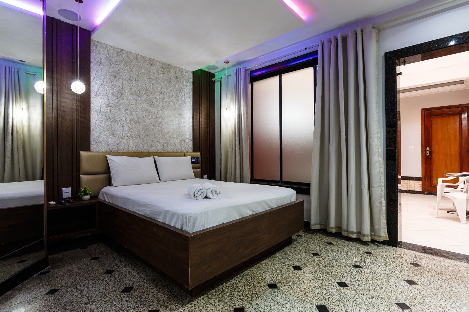 Quarto com cama de casal, cortinas bege e piso estampado. Detalhes em madeira escura e luminárias coloridas no teto.