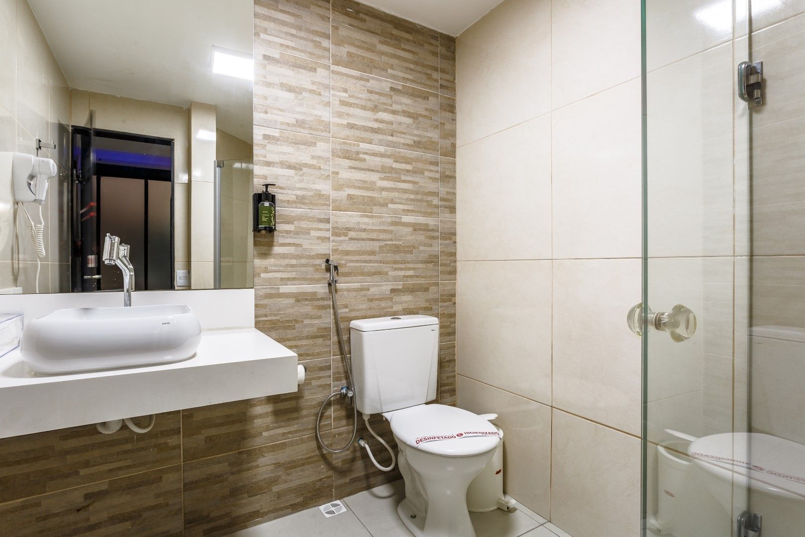 Banheiro com acessórios brancos, azulejos bege e box de vidro.