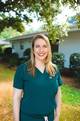 Woman standing outdoors in a green shirt, smiling in front of a house and trees.