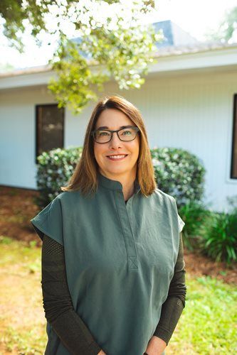 Smiling person in glasses standing outdoors in front of a white building and greenery