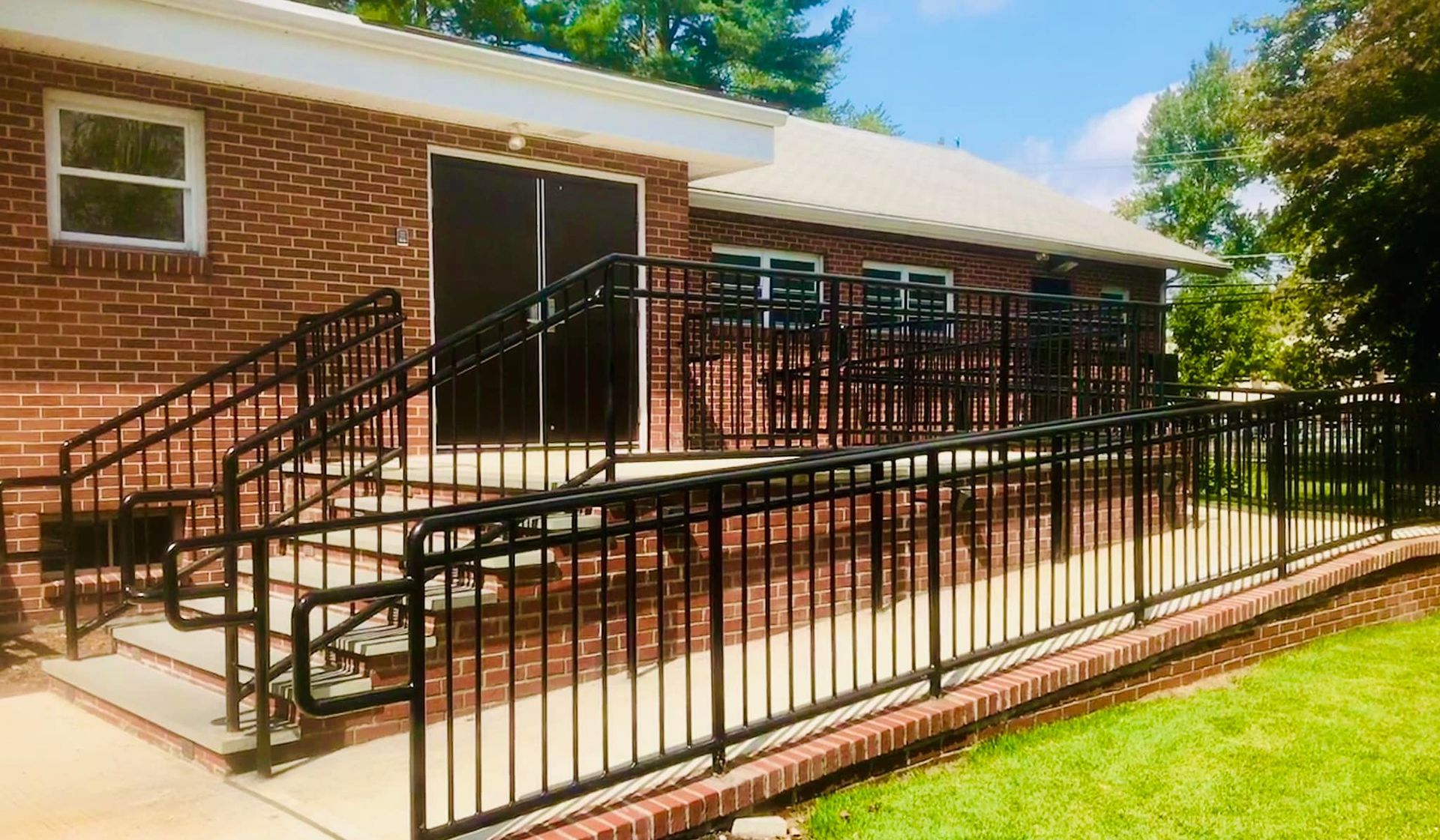 Brick building entrance with black metal ramps and railings, leading to a patio and lawn.