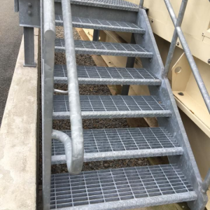 Metal staircase with grated steps and a handrail beside a beige industrial wall.