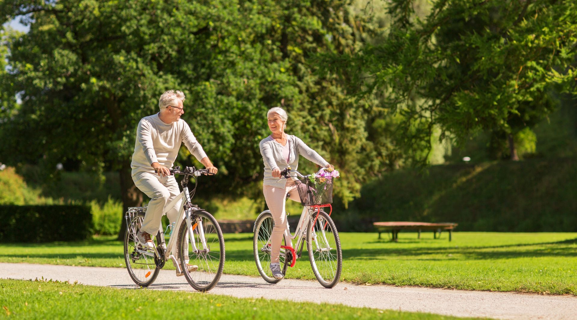 Zwei ältere Frauen fahren mit dem Fahrrad auf einem gepflasterten Weg in einem sonnigen Park, grünes Gras und Bäume im Hintergrund.