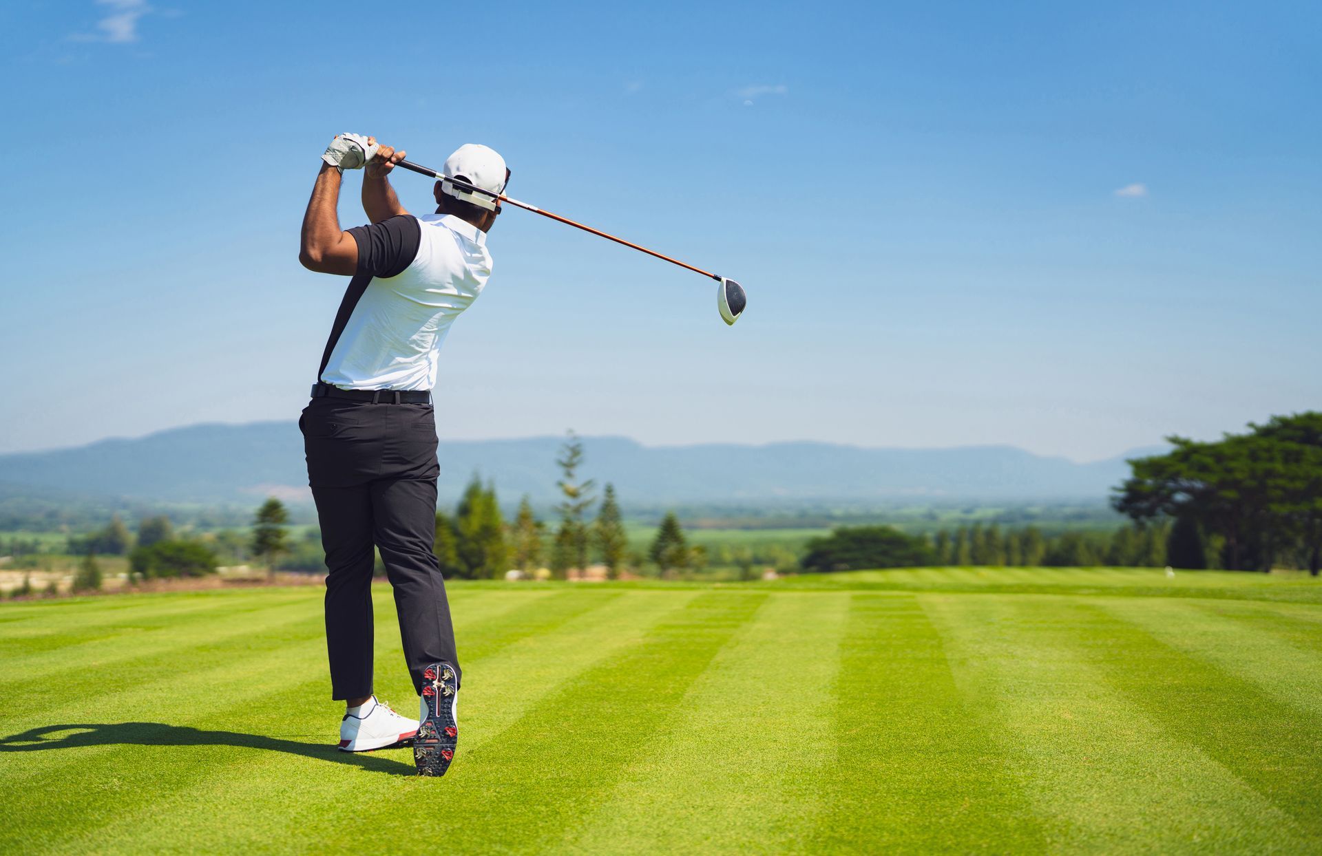 Golfer schwingt Schläger auf Grün, blauer Himmel, Berge im Hintergrund.