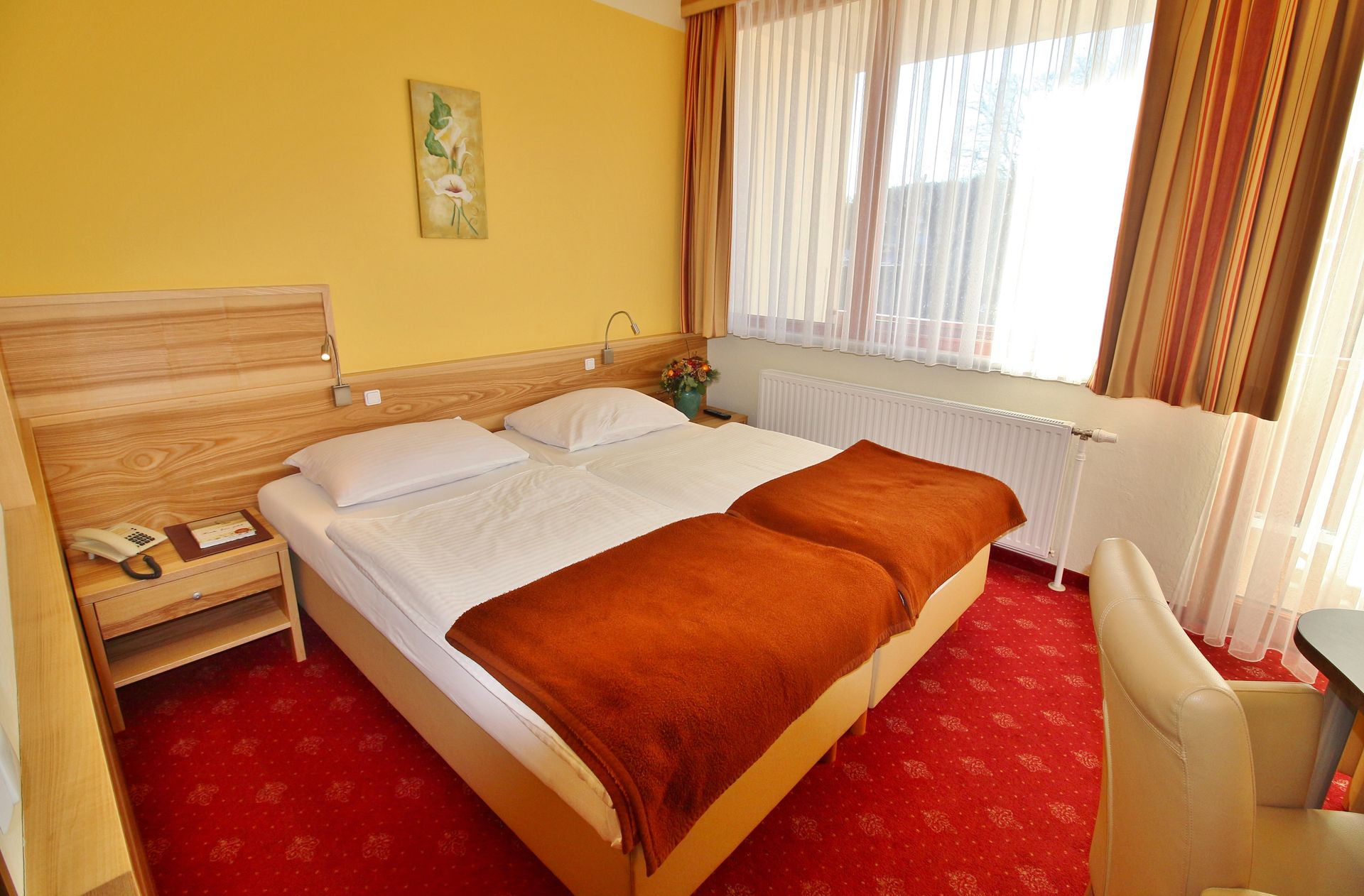 Hotelzimmer mit Doppelbett, rotem Teppich, gelben Wänden und einem Fenster mit Vorhängen.