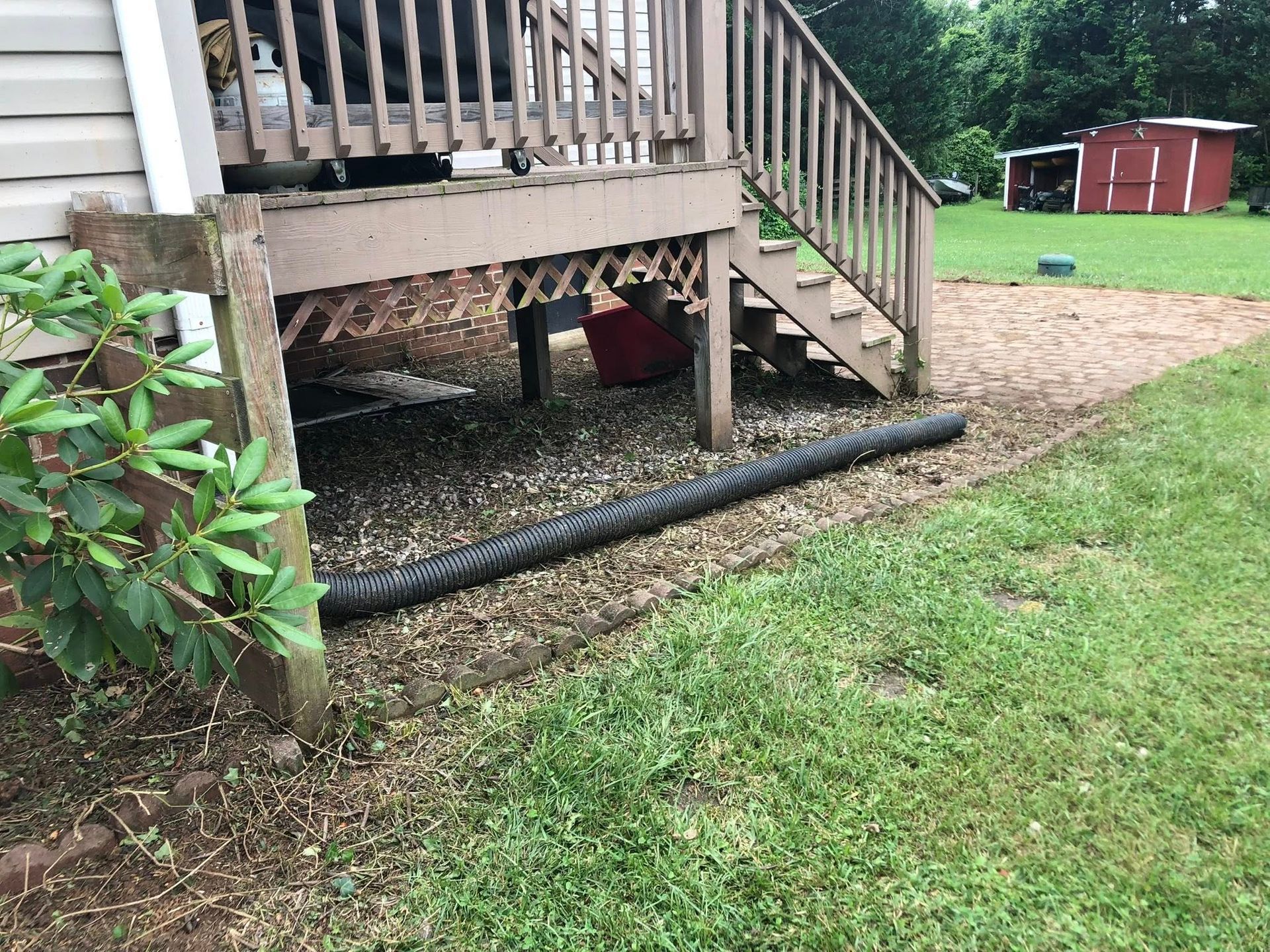 Under deck with gravel, drainage pipe, wooden stairs and shed in background. Green grass.
