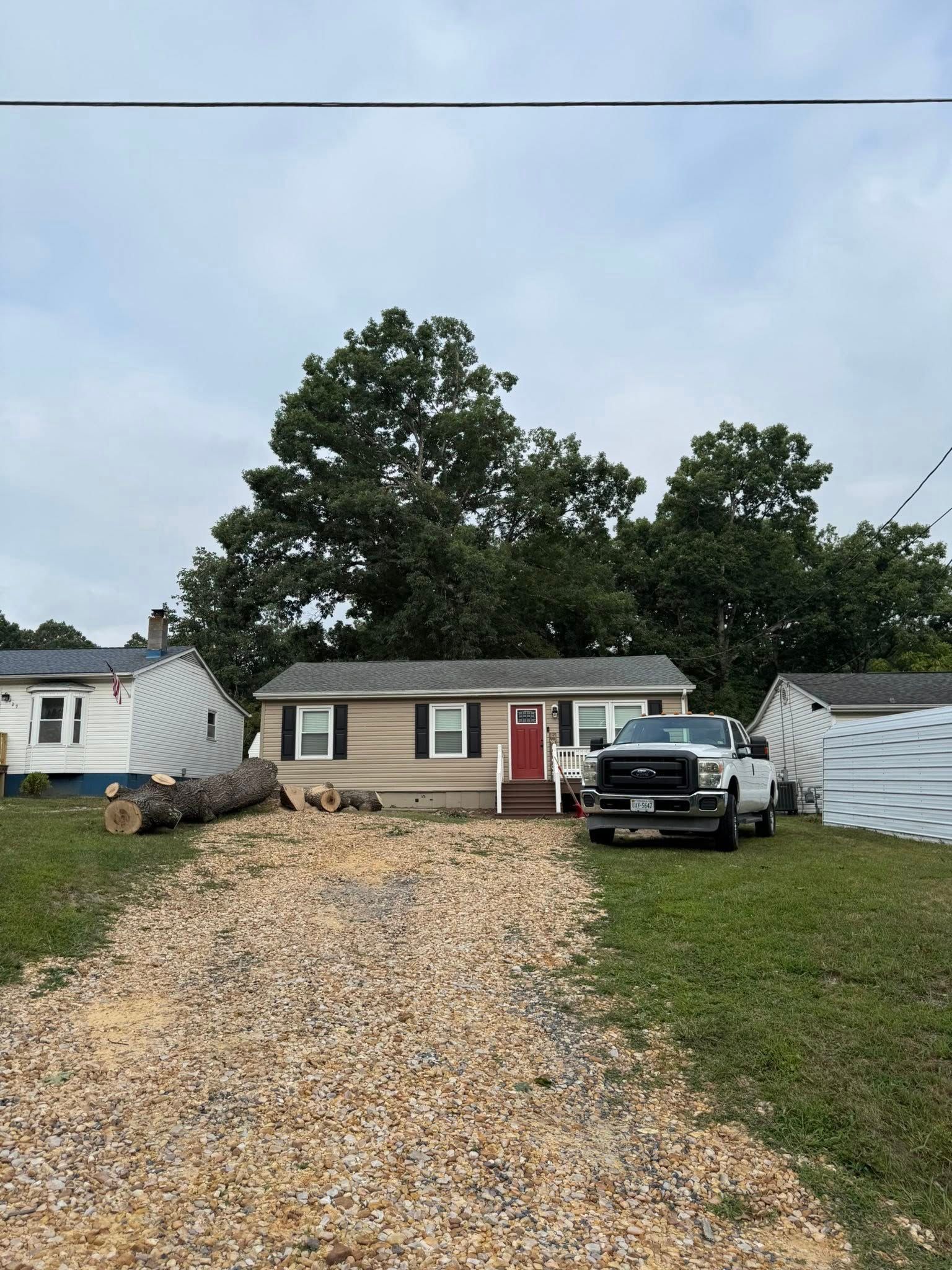 A small, tan house with a red door has a gravel driveway. A pickup truck is parked in front.