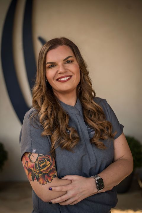 A woman in a purple scrub top is smiling with her arms crossed.