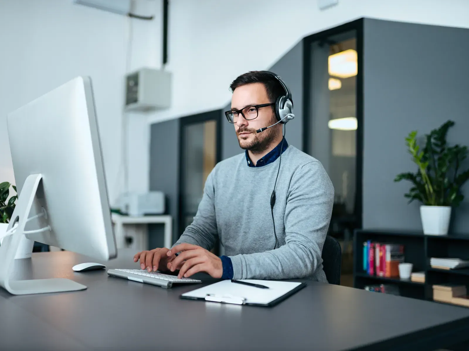 Man wearing a headset typing at a desk in a modern office with a large monitor